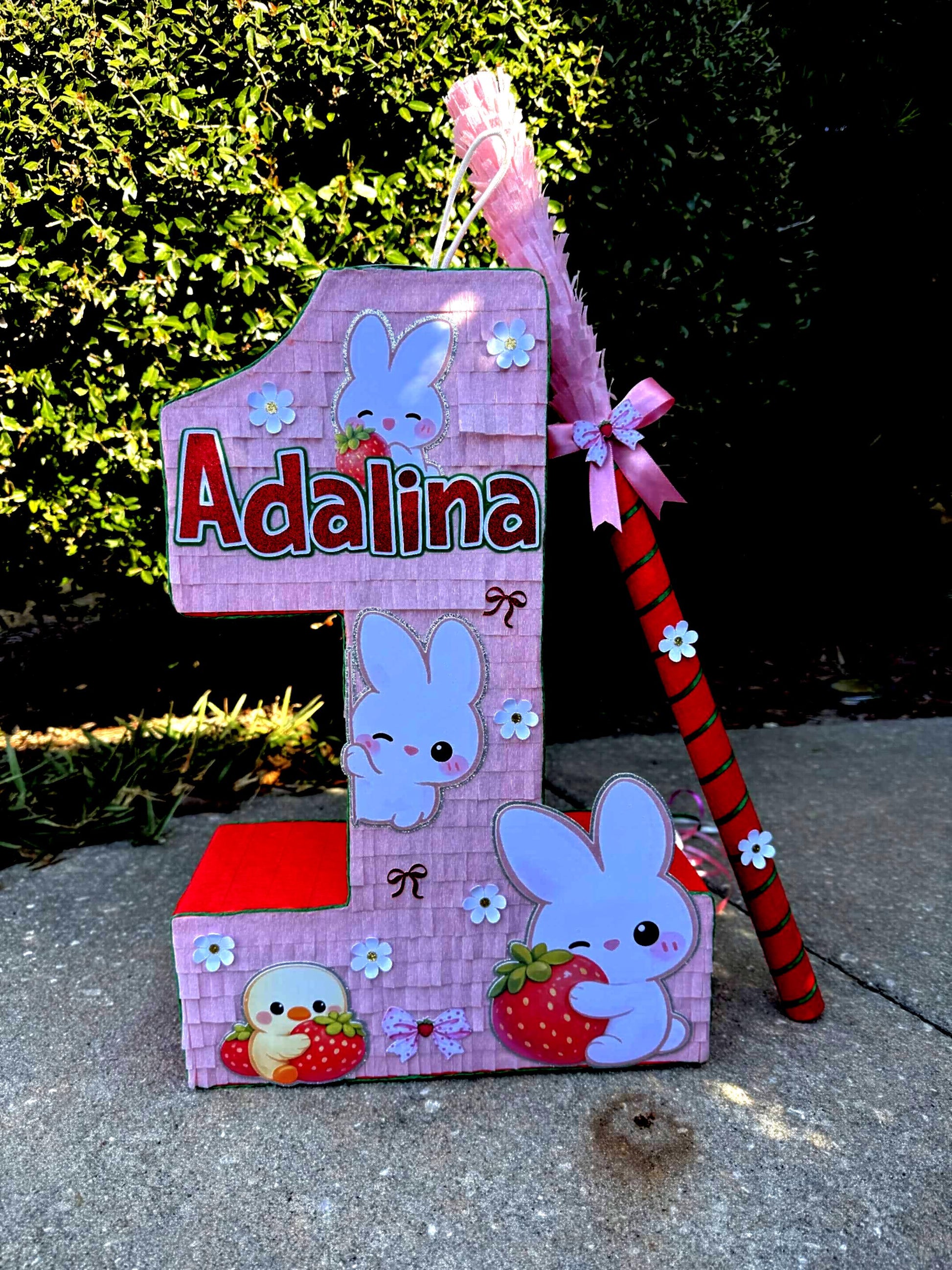 May include: A pink number one piñata with the name "Adalina" in red letters. The piñata is decorated with white cartoon bunnies, strawberries, and small white flowers. A pink and red stick is included.